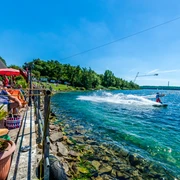 Wasserskianlage am Kulkwitzer See - Leipziger Neuseenland Eine Person fährt mit einem Wakeboard über das kristallklare Wasser des Kulkwitzer Sees, während Besucher auf einer sonnigen Terrasse entspannen.
