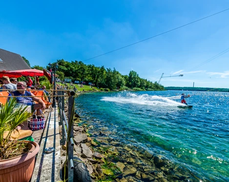 Wasserskianlage am Kulkwitzer See - Leipziger Neuseenland Eine Person fährt mit einem Wakeboard über das kristallklare Wasser des Kulkwitzer Sees, während Besucher auf einer sonnigen Terrasse entspannen.
