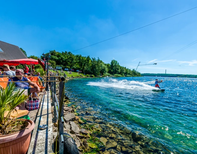 Wasserskianlage am Kulkwitzer See - Leipziger Neuseenland Eine Person fährt mit einem Wakeboard über das kristallklare Wasser des Kulkwitzer Sees, während Besucher auf einer sonnigen Terrasse entspannen.