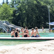 NaturSportBad Bad Düben - Freibäder in der Leipzig Region Sechs Kinder sitzen am Rande des Beckens mit dem Rücken zur Kamera in Badesachen und lassen ihre Beine im Wasser baumeln, im Hintergrund ist eine große Rutsche zu sehen, Freizeit, Spaß
