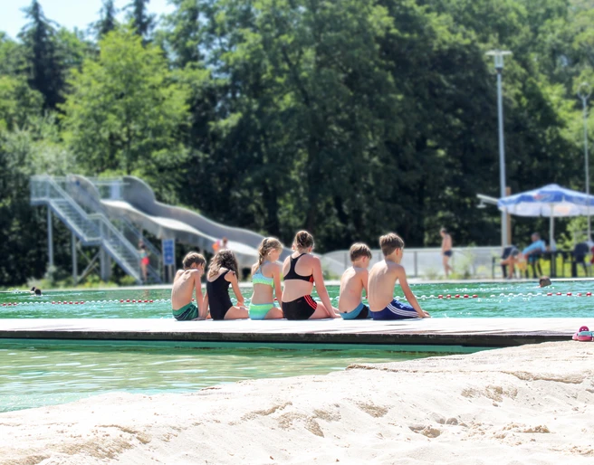 NaturSportBad Bad Düben - Freibäder in der Leipzig Region Sechs Kinder sitzen am Rande des Beckens mit dem Rücken zur Kamera in Badesachen und lassen ihre Beine im Wasser baumeln, im Hintergrund ist eine große Rutsche zu sehen, Freizeit, Spaß