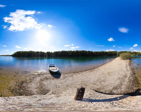 Albrechtshainer See im Leipziger Neuseenland Das Bild zeigt einen ruhigen See mit klarem Wasser, das sanft ans Ufer schwappt. Am linken Bildrand liegt ein bescheidenes Boot, bereit zur Abfahrt. Die Szenerie wird von einem strahlend blauen Himmel mit wenigen weißen Wolken überdeckt.