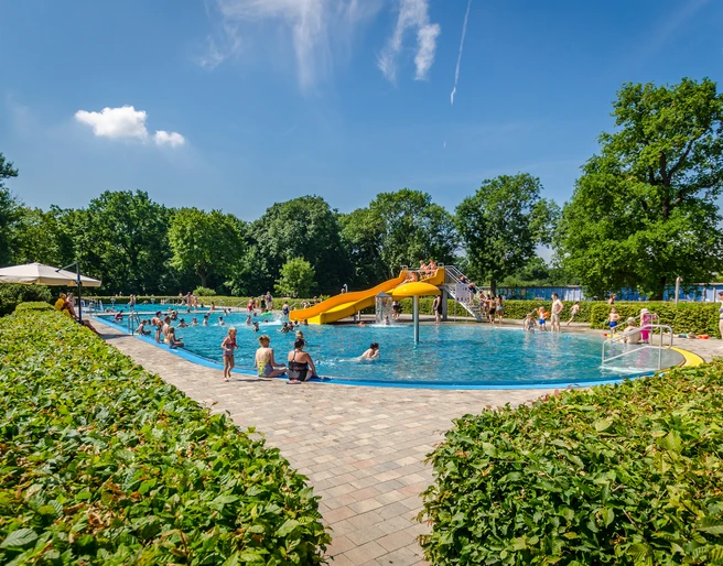 Schreberbad Leipzig - Freizeiterlebnisse in der Leipzig Region Blick auf das Hauptschwimmbecken im Schreberbad, mit gelber Rutsche und vielen Badegästen an einem Sommertag, Freizeitaktivität, Badespaß