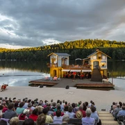 Seebühne Kriebstein - Kultur in Leipzig und Region Foto von der Seebühne Kriebstein auf der Talsperre Kriebstein bei Sonnenuntergang, auf der Bühne ist eine Kulisse mit 2 Häusern aufgebaut und in der Mitte spielt ein Orchester, Veranstaltung