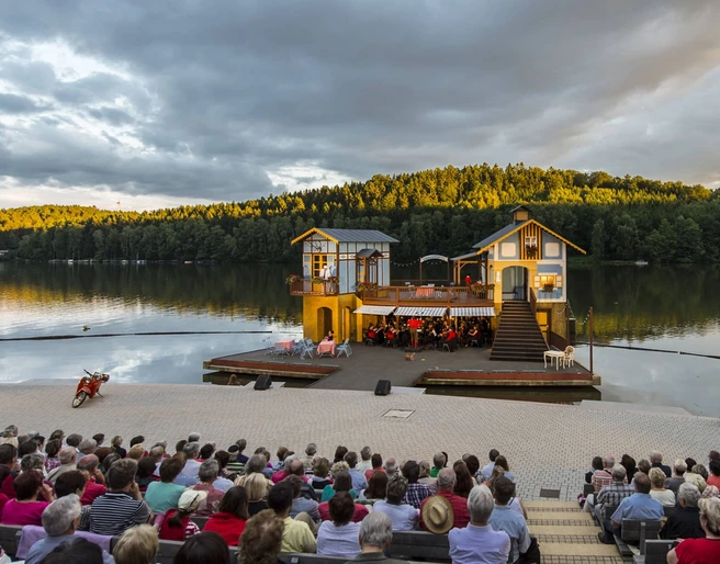 Seebühne Kriebstein - Kultur in Leipzig und Region Foto von der Seebühne Kriebstein auf der Talsperre Kriebstein bei Sonnenuntergang, auf der Bühne ist eine Kulisse mit 2 Häusern aufgebaut und in der Mitte spielt ein Orchester, Veranstaltung