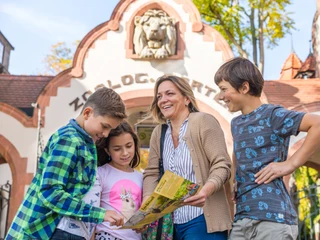 Zoo Leipzig - Leipzig mit Kindern Eine Familie steht vor dem Eingangsportal des Zoo Leipzig und schaut in den Faltplan des Zoo Leipzig, Familie, mit Kindern, Freizeit, AusflugA family stands at Leipzig Zoo's entrance portal while browsing the zoo brochure, family, children, leisure, outingsUne famille devant l’entrée du zoo de Leipzig regarde le plan dépliant du zoo de Leipzig, famille, avec enfants, loisirs, excursionRodina stojí před vchodem do Lipské zoo a dívá se na skládací mapu Lipské zoo, rodina, s dětmi, volný čas, výletRodzina stoi przed bramą lipskiego zoo i ogląda mapkę obiektu, rodzina, z dziećmi, czas wolny, wycieczka