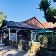 Eingang Restraurant Restaurantfront mit roten Ziegeln, weißen Holzleisten und einem Garten mit Sonnenschirm.