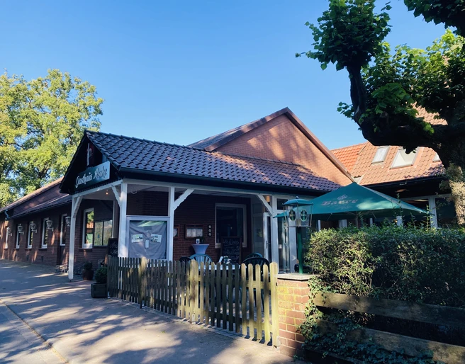 Eingang Restraurant Restaurantfront mit roten Ziegeln, weißen Holzleisten und einem Garten mit Sonnenschirm.