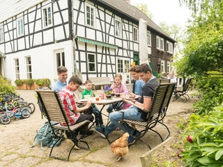 Pause im Hofcafé auf dem Hof zur Hellen im Windrather Tal Menschen sitzen an einem Tisch im Freien eines Fachwerkhauses. Ein Huhn spaziert um den Tisch.