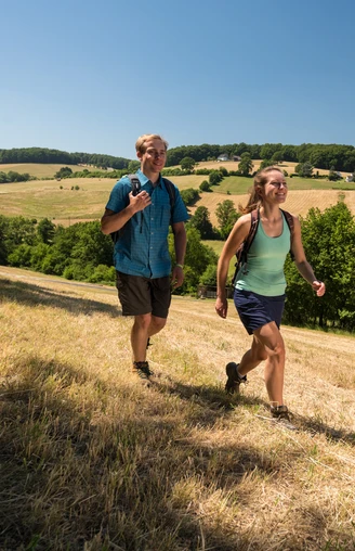 Wandergenuss in der Elfringhauser Schweiz Zwei Personen wandern auf einem grasbewachsenen Hügel mit weiten Feldern und Bäumen im Hintergrund.