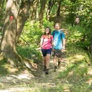 Unterwegs auf schmalen Pfaden durch verwunschen wirkende Wälder Zwei Personen spazieren auf einem schmalen Waldpfad, umgeben von dichtem Grün und Bäumen.