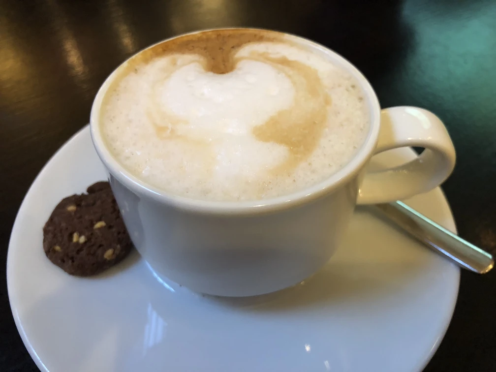 Kaffeetasse Eine weiße Tasse mit aufgeschäumtem Cappuccino, serviert auf einem weißen Unterteller neben einem Schokoladenkeks.