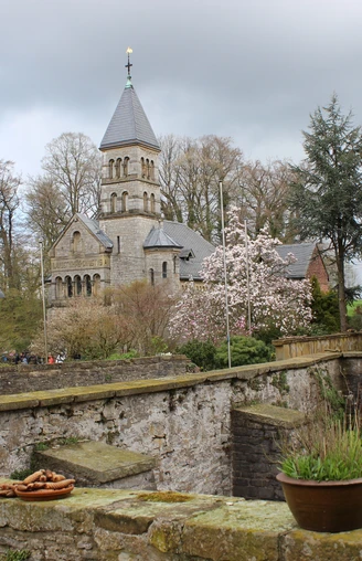 Historische Kirche mit spitzem Turm, blühende Bäume und alte Mauer in malerischer Frühlingslandschaft.