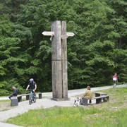 Ein hölzernes Wanderkreuz mit Wegweisern steht vor grünen Bäumen, umgeben von Radfahrern und Sitzbänken.
