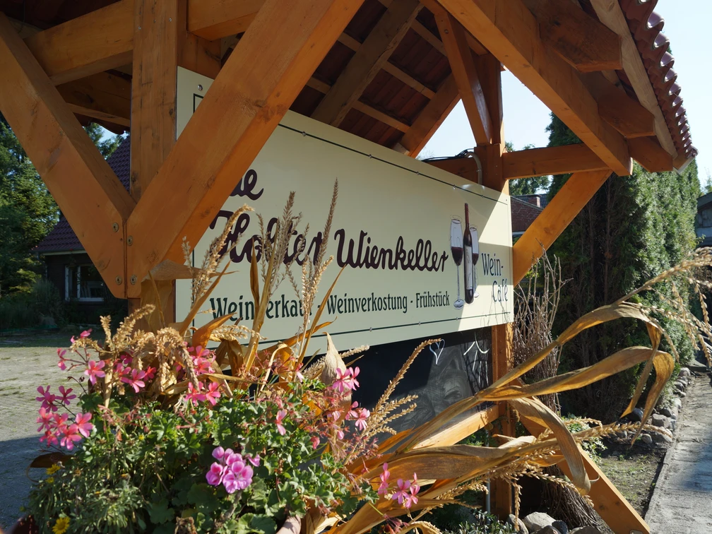 De Holter Wienkeller in Holte, Gemeinde Rhauderfehn, DSC01033.JPG Holzunterstand mit Schild für Weingut Ulienkeller, davor Blumenkasten mit pinken Blüten und getrockneten Gräsern.