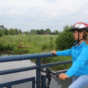 Meliorationstour Radfahrerin mit Schutzhelm betrachtet von einer Brücke einen ruhigen Flusslauf inmitten grüner Natur.
