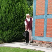 Eine Frau in historischer Tracht blickt lächelnd hinter einer Fachwerkfassade hervor, umgeben von Grün.