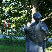 Der Zeitungsleser Steinskulptur eines Menschen mit Hut, der eine Zeitung hält, umgeben von Grün und Bäumen.
