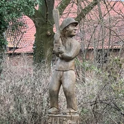 Holzskulptur eines Schlagballspielers im Wald, zeigt feste Haltung und traditionellen Spielstil.