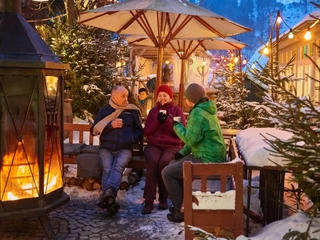Glühweinplausch im Mühlenhof  Drei Personen sitzen in winterlicher Atmosphäre um eine Feuerstelle, umgeben von schneebedeckten Tannen und bunten Lichtern, und trinken heiße Getränke.