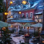Winterdorf Schmilka Mühlenhof Verschneiter Innenhof mit beleuchteten Sternen an Bäumen, Holzbänken, grünen Sonnenschirmen und Laternen, umgeben von Fachwerkhäusern.