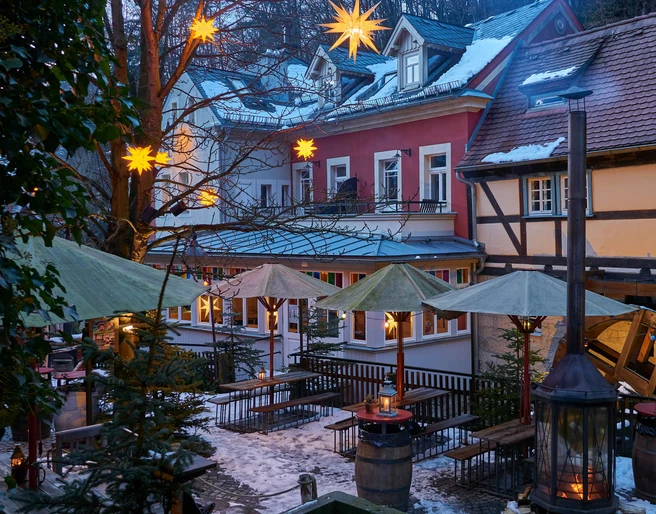Winterdorf Schmilka Mühlenhof Verschneiter Innenhof mit beleuchteten Sternen an Bäumen, Holzbänken, grünen Sonnenschirmen und Laternen, umgeben von Fachwerkhäusern.