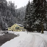 Gasthaus Polenztal Gelbes Gebäude in verschneiter Waldlandschaft, neben einem Bach und schneebedecktem Weg, umgeben von hohen Tannenbäumen.