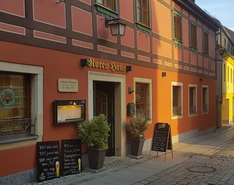 Gasthaus "Zum Roten Haus" Fachwerkhaus mit roter Fassade und grünen Fensterläden, beschriftet als "Rotes Haus", mit Menütafeln und Pflanzen vor der Tür.