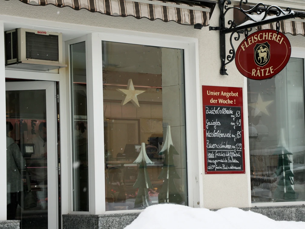 Eingang einer Fleischerei mit rotem Schild und Wochenangebot auf einer Tafel; Fenster mit Stern- und Tannenbaumdekoration, Schnee im Vordergrund.