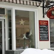 Fleischerei Rätze Eingang einer Fleischerei mit rotem Schild und Wochenangebot auf einer Tafel; Fenster mit Stern- und Tannenbaumdekoration, Schnee im Vordergrund.
