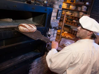 Holzofen in der Bio-Bäckerei in Schmilka Bäcker in weißer Kleidung zieht frisches Brot mit einer Schaufel aus einem traditionellen Backofen; im Hintergrund Regale mit weiteren Broten.