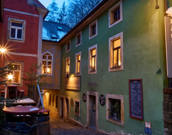 Bio-Bäckerei im Winter Enge Gasse mit einer rot-grünen Bäckerei, beleuchtet von warmem Licht, weihnachtliche Dekorationen in den Fenstern, winterliche Atmosphäre.