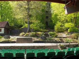 Freilichtbühne Nettelstedt.jpg Grüne Sitzreihe vor Freilichtbühne im Wald mit zwei Häuschen und Türmen, umgeben von Bäumen.