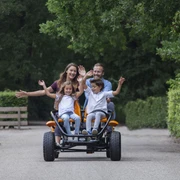 Gocart Giebner in Monheim am Rhein Eine Familie fährt auf einem viersitzigen, orangenen Gocart fröhlich auf einer von Bäumen gesäumten Straße.