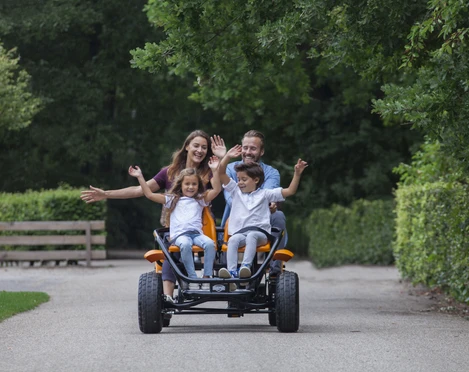 Gocart Giebner in Monheim am Rhein Eine Familie fährt auf einem viersitzigen, orangenen Gocart fröhlich auf einer von Bäumen gesäumten Straße.