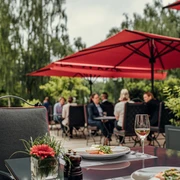 Auf der Außenterrasse des Bistrots Quellenhof befinden sich mehrere Sitzgruppen mit Personen. Sie werden von roten, quadratischen Schirmen überdacht. Im Vordergrund steht ein mit Essen und Gläsern gedeckter Tisch und ein leerer Stoffstuhl.Im Hintergrund stehen Bäume.