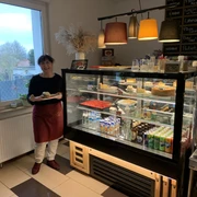 Auslage in einem Café. Man sieht eine Frau, die einen Teller mit einem belegten Brötchen hält. Neben ihr ist eine Vitrine mit Kuchen, Limonade und Säften.