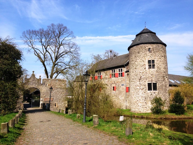 Wasserburg Haus zum Haus in Ratingen Wasserburg mit rundem Turm, von Bäumen umgeben, blauer Himmel und Kopfsteinpflasterweg im Vordergrund.