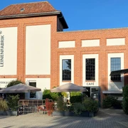 LEINENFABRIK Außenansicht Gebäude Backsteingebäude mit grauer Fassade, schattiger Terrasse, Verkaufsraum und Café unter Sonnenschirmen.