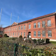 Backsteinbau der Polizeiakademie mit großen Fenstern, umgeben von gepflegten Pflanzen und leeren Fahnenmasten.