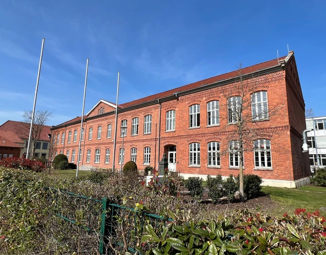Polizeiakademie Backsteinbau der Polizeiakademie mit großen Fenstern, umgeben von gepflegten Pflanzen und leeren Fahnenmasten.