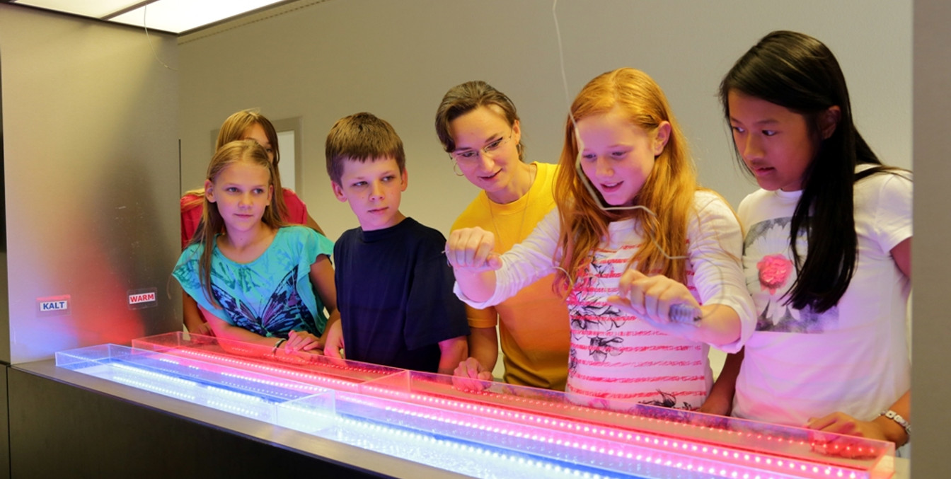 4 Kinder stehen vor einem Experiment, ein Mädchen hält einen Draht in der Hand und alle anderen Personen schauen gespannt auf sie, Freizeit-Museum-Leipzig-mit-Kindern