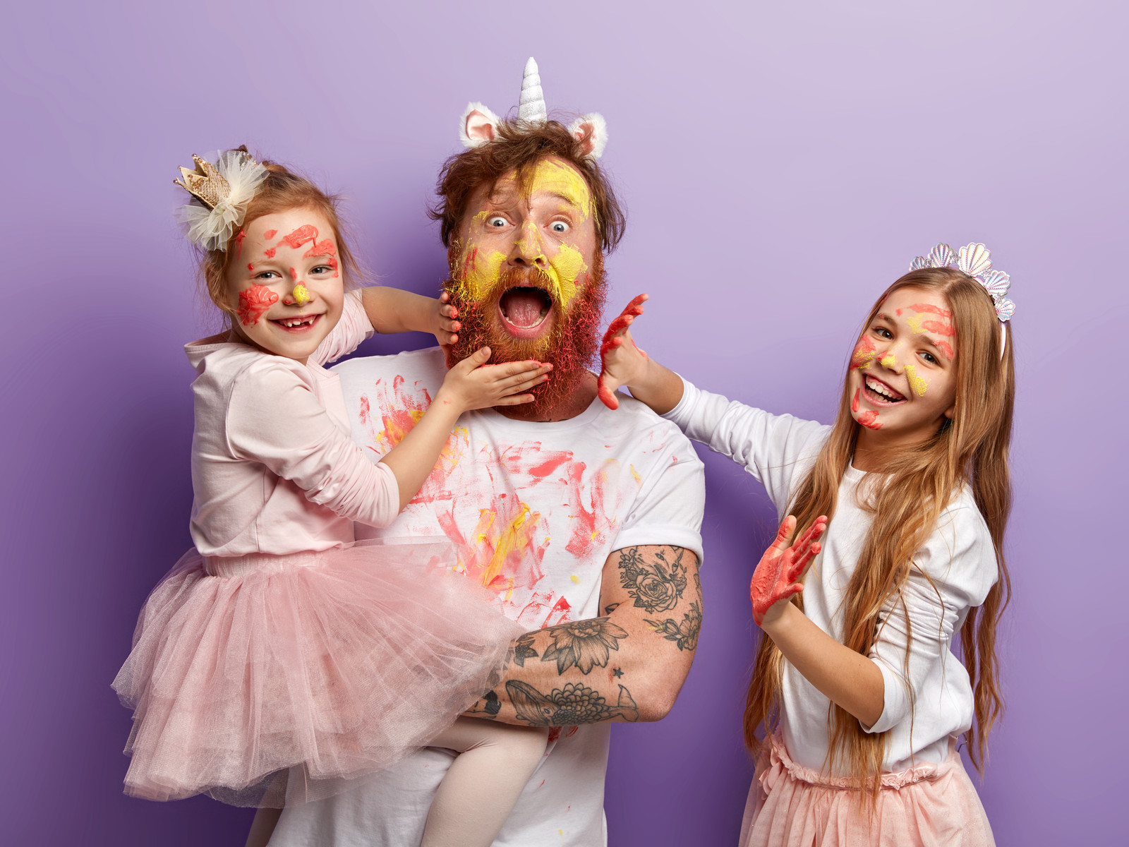 Emotional bearded busy father spends time with two naughty daughters who leave palm prints on his beard and clothes, learn how to paint, dressed in festive clothes, stand indoor. So colourful!
