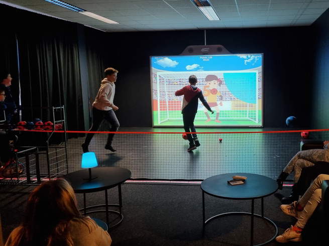 Zwei Kinder spielen an der XXL Gamingwand von Team-Duell Leipzig Elfmeter, währendessen es sich die Zuschauer in der Lounge-Area bequem machen und den Spielspaß beobachten