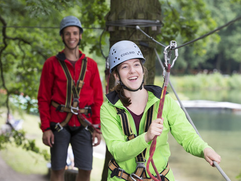 Eine Frau in Kletterausrüstung klettert an einem Seil entlang, hinter ihr steht ein Mann an einem Baum.A woman in climbing gear is climbing along a rope, behind her is a man standing against a tree.