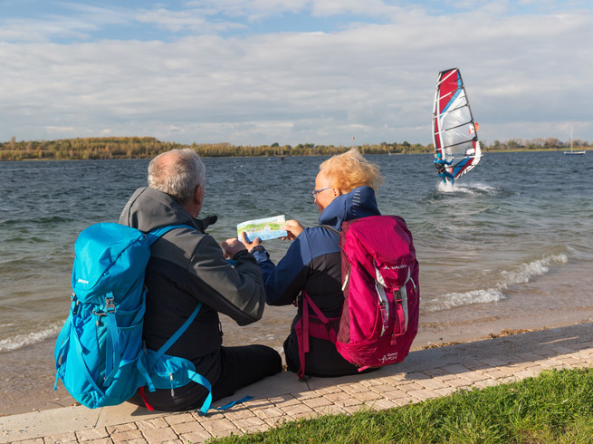 Rückansicht eines älteren Paares, das sich am Rande des Schladitzer Sees ein Foto anschaut, Surfer, Boote, Wälder, blauer Himmel