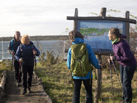 Vier Wanderer begegnen sich auf einem Wanderweg, daneben eine Landkarte, im Hintergrund der Schladitzer See