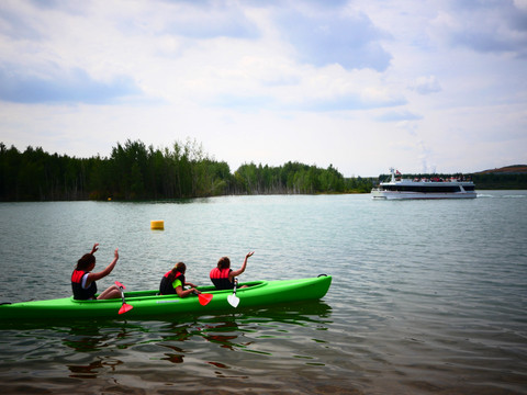 Drei Frauen sitzen in einem Kanu auf dem Markkleeberger See und winken einem vorbeifahrendem Fahrgastschiff, Leipziger Neuseenland, Wassersport, Freizeit