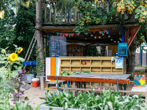 Blick auf den Annalinde-Kiosk im Garten, seine Angebotstafel und die vielen Blumen und Pflanzen um den Gartenverschlag herum, Urbane Landwirtschaft, Gastronomie, Bar, Natur, Freizeit