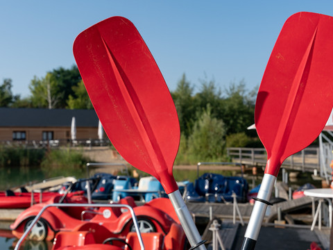Zwei rote Paddel kreuzen sich, im Hintergrund Tretboote auf dem Verleihgelände, leipziger neuseenland, aktiv, wassersport